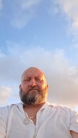A man with a shaved head and a beard is looking at the camera. He is wearing a white shirt with a pattern on it and a gold chain with a Star of David pendant. The sky behind him is blue with clouds, and the sun is setting, casting a warm glow on the clouds.