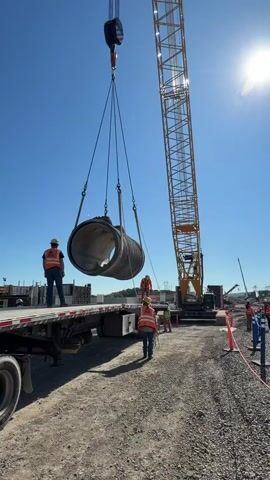 The video captures a daytime scene at a construction site in Center Township, United States of America, under a clear, sunny sky. A large lattice boom crane is actively lifting a substantial concrete pipe, suspended by slings from its hook, off the back of a flatbed trailer.

Three construction workers are visible, all wearing safety vests and hard hats. One worker stands on the flatbed trailer, observing the lift. Another worker is on the dusty ground near the suspended pipe, and a third is further back near the base of the crane. The sun casts strong shadows, indicating bright conditions. In the background, other construction equipment, materials, and power lines or towers are discernible. The word "BENSON" is visible in black lettering on the side of the flatbed trailer. The overall activity appears to be the handling of large infrastructure components for a construction project.