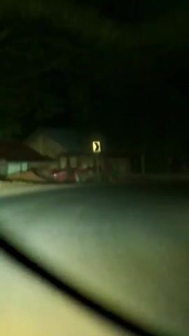 The video begins with a blurred view of a dark, tree-lined road at night. A yellow directional sign indicating a curve to the right is visible on the left side of the road. In the distance, a building with lights on appears to be on the left. A red object, possibly a vehicle, is parked in front of it. As the video progresses, the perspective shifts, showing the road and trees passing by. The headlights of another vehicle are visible in the distance. The scene is predominantly dark with the ambient light of the headlights and streetlights illuminating the surroundings.
