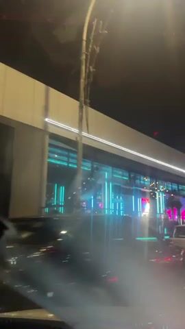 The video captures a nighttime street view of a modern building with bright neon blue and pink lights. A sign on the building reads "rumours.". Cars are parked and driving in front of the building, and the view appears to be from inside another vehicle, as indicated by reflections on the glass. The lighting suggests a lively urban environment, possibly a nightlife district.