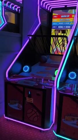 The video shows a row of arcade games with neon lighting. The games appear to be basketball themed and Connect 4. The games are lit with blue and red neon lights. The camera pans across the games, showing different angles of the machines. The overall scene is vibrant and energetic, typical of an arcade environment.