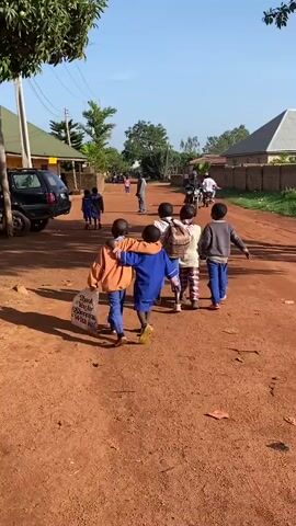 The video captures a daytime street scene in Kuru, Nigeria. The primary subjects are a group of children walking away from the camera along a reddish-brown dirt road. In the foreground, two boys are seen walking arm-in-arm, one in a blue top and the other in an orange jacket. Other children walk alongside them; one child carries a white plastic bag.

Further down the road, more people are visible, including other children and adults. Some individuals are walking, while others are riding motorcycles. On the left side of the road, a black SUV is parked near a building, partially obscured by trees. Residential buildings with varying roof colors (green, gray) line both sides of the road. The setting suggests a semi-urban or rural residential area.

The weather is bright and sunny, indicated by the clear blue sky and visible shadows on the ground. Trees and bushes provide vegetation throughout the scene. The activity depicts typical daily movement within a community. No visible text is clearly legible in the video.