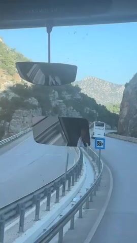 The video shows a view from inside a vehicle traveling on a highway. The road curves to the right, with a steep, rocky, and tree-covered mountainside on the left. A white bus and a car are visible ahead on the road. A metal guardrail runs along the left side of the road. In the foreground, a rearview mirror reflects a person wearing a light-colored shirt. Another, smaller rearview mirror is positioned above it. A blue sign with a camera icon is visible on the left side of the road. The sky is clear and blue.