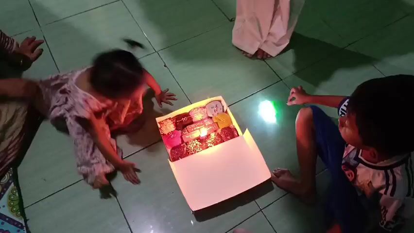 A group of people are gathered around a box of donuts with a lit candle. Two young children are sitting on the floor, one on the left facing the box and the other on the right facing away from the box. An adult woman in a hijab is kneeling on the right, holding a smartphone and a flashlight. Another adult, partially visible on the left, has their hand near the box. The scene appears to be indoors, with a tiled floor and dimly lit surroundings. The atmosphere suggests a celebration, possibly a birthday.
