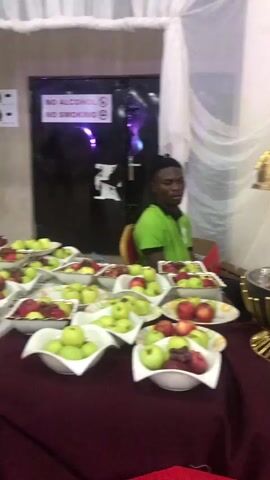 A man in a green shirt sits behind a table laden with plates of apples and grapes and a gold-colored punch bowl. The camera moves from left to right, showing the full table, where several of the same bowls and fruit plates are set out. Behind the table is a white curtain. A man in an orange shirt stands to the right of the frame. The camera zooms out to show the decorations and layout of the room, including white chairs and tables. The overall impression is of a buffet at an event.
