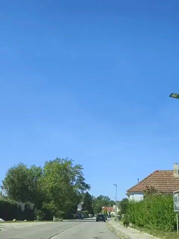 This is a first-person perspective video taken from a vehicle driving down a paved road in a suburban area. The sky is clear and blue. On the left side of the road, there are trees and a white fence. On the right side, there are houses with red-tiled roofs and green hedges. There are several cars on the road, moving in the same direction as the vehicle from which the video is being filmed. The video shows a peaceful daytime scene in a residential neighborhood.
