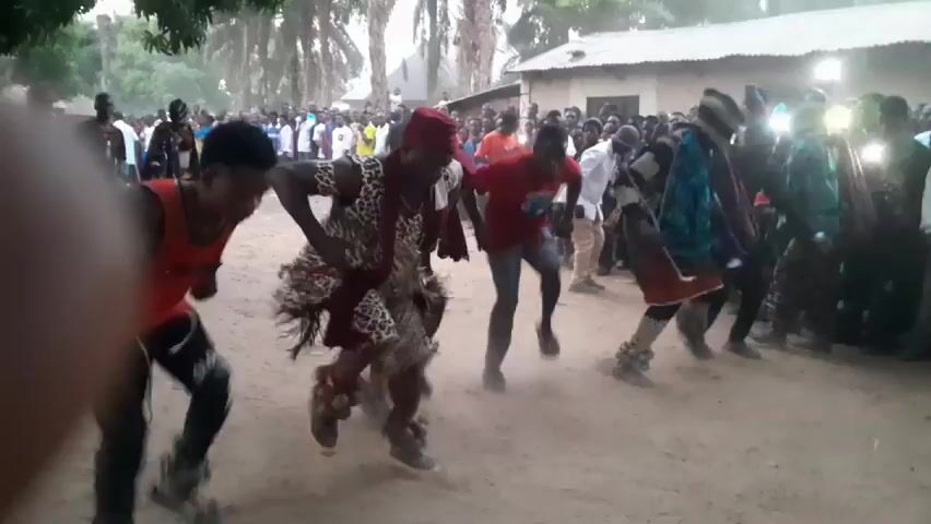 A group of people are dancing outdoors in what appears to be a village setting. Dust rises from the ground as they move. Several individuals are wearing traditional clothing or costumes, including animal prints and fringed garments. The crowd is a mix of ages and genders. Some people in the background are holding up their phones to record the event. The setting appears to be a dusty open area with some trees and buildings in the background.