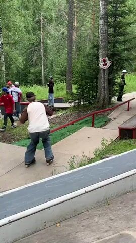A young person on rollerblades is grinding down a red metal rail on an outdoor skate park. The person is wearing a beige t-shirt, blue jeans, a black baseball cap, and black rollerblades. The rail is positioned over a concrete ramp covered with green turf. In the background, there are trees and other skate park obstacles. Another person, wearing a helmet and camouflage clothing, stands near the end of the rail, observing. A third person in a black shirt and blue jeans watches from further back, near a wooden structure. The weather appears to be overcast.