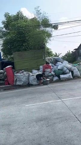 The video shows a roadside scene with several large, white sacks piled up, some with red bags. A person in a black shirt and shorts is sorting through some of the sacks. A green corrugated metal structure is behind the sacks. Trees and power lines are visible in the background. Later, a person on a motorcycle with two passengers rides past the scene. The weather appears to be partly cloudy.