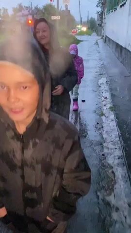 The video shows two people walking on a wet path in what appears to be a rural or suburban area during rainfall. The first person is partially visible in the foreground, wearing a dark hooded jacket. The second person is further ahead and in focus, wearing a dark hooded coat and smiling. The path is paved and wet, with puddles reflecting the dim, overcast sky. To the right of the path is the side of a building with a textured facade. To the left, there are some grassy areas and more buildings in the distance. The video then cuts to a child wearing a bright pink hooded jacket and pink rain boots, walking on the same wet path. The child is facing the camera and appears to be smiling. Another person, wearing a pink hooded garment, is partially visible in the foreground of the last clip, walking away from the camera. The overall environment suggests a rainy day with poor visibility due to the weather.