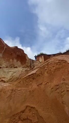 A low-angle, vertical shot captures a person standing on the edge of a sandy, orange-toned cliff. The person is wearing a dark t-shirt and blue jeans. They are facing forward with their hands on their hips. The cliff face is striated with various shades of red and tan rock, suggesting layers of sediment. The sky above is blue with scattered white clouds. The ground in the foreground is also made of the same sandy, orange material, appearing eroded and textured. The overall scene depicts a natural outdoor environment, possibly a canyon or a sand pit.