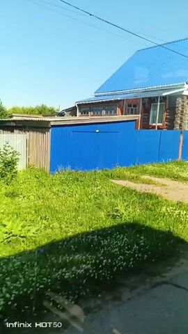 The video shows a view from a vehicle moving along a street. The scene is a rural area with a wooden house with a blue roof, a blue fence, and green grass. Trees are visible in the background. The weather appears to be sunny with a clear blue sky. The video has a timestamp of 00:00.