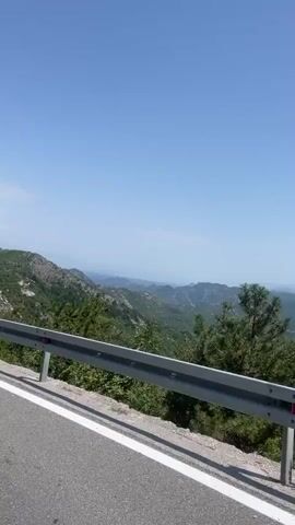This video captures a scenic drive through a mountainous region, likely in Dobrsko selo, Montenegro. The camera, mounted from a vehicle, offers a view over a guardrail, revealing a vast landscape of rolling green hills and distant mountains under a clear blue sky. The sun appears high, suggesting midday. The scene is dominated by nature, with lush vegetation covering the slopes, interspersed with rocky outcrops and scattered trees.

The primary activity is travel by road. The highway, with white lane markings, curves gently, leading the viewer deeper into the vista. Several vehicles are visible on the road ahead, including a dark-colored sedan and a few other cars, indicating moderate traffic. The presence of a guardrail along the road's edge is a notable safety feature. In the foreground, a motorcycle's rearview mirror is visible, reflecting a person's face wearing a white shirt, suggesting the video is from the perspective of a motorcyclist.

The overall mood is serene and adventurous, highlighting the beauty of the natural environment and the freedom of the open road. The weather is fair and sunny, ideal for outdoor exploration. The landscape's ruggedness and greenery point towards a natural, possibly rural or mountainous, setting. No visible text is present in the video.