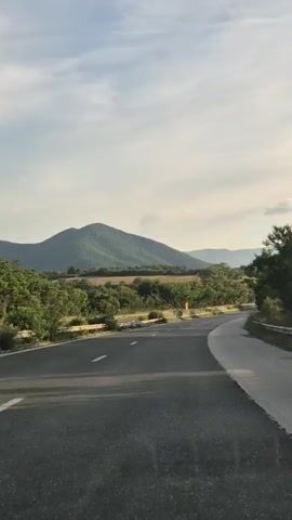 The video shows a paved road curving to the right, with a white line marking the left lane and a white border marking the right lane. The road is bordered by a guardrail on the left and trees on the right. In the distance, there are green hills and mountains under a sky with scattered clouds. The scene appears to be a daytime shot, likely from a moving vehicle.