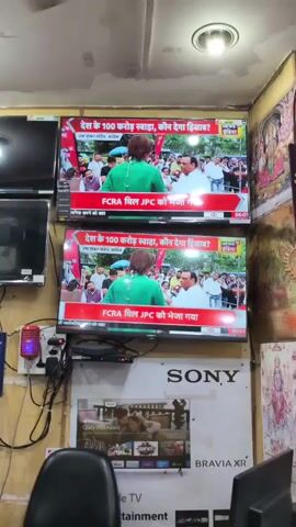 The video shows two identical television screens displaying a news report in Hindi. Both screens show a woman in a green top facing away from the camera, with her arms crossed. She is standing in front of a crowd of people. A man in a white shirt is standing to her right. The text on the screen reads "देश के 100 करोड़ स्वाहा, कौन देगा हिसाब?" and "FCRA बिल JPC को भेजा गया". Below the televisions, a Sony Bravia XR advertisement is visible. The environment appears to be an electronics store, with other televisions and advertisements in the background. The lighting is overhead, and there is a purple lamp visible on the left.