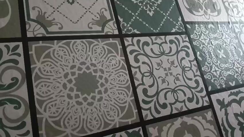 This video shows a close-up view of a patterned fabric or surface, possibly a tablecloth or upholstery. The pattern consists of repeating square tiles, each featuring a different intricate design in shades of green and grey on a white background. The lighting suggests a bright, possibly natural, light source from the right side of the frame, creating some highlights and shadows across the surface. The camera is slightly tilted and moves slowly across the pattern, allowing a detailed examination of the geometric and floral motifs within the tiles. There are no people or discernible actions taking place, the focus is solely on the static visual pattern.