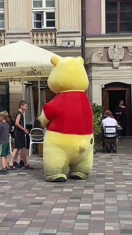 A large, inflatable Winnie the Pooh costume character stands in a cobblestone plaza. The character is facing away from the camera, with its back to the viewer. Several people are visible in the background, including a group of children and adults gathered near the character, some sitting at outdoor tables under a white awning. The plaza appears to be in a European city, with a building featuring classical architecture in the background. The character appears to be interacting with the children.