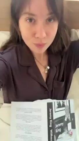 A woman with long, dark brown hair and bangs is lying in bed, holding open a book titled "Jeremy King Without End." She is wearing a dark-colored, long-sleeved pajama top, small hoop earrings, and a necklace with charms. She has a small tattoo of a lightning bolt on her left wrist. The woman looks at the camera, touches her hair, and rests her face in her hand.  Behind her, a window is visible with framed artwork on the wall.