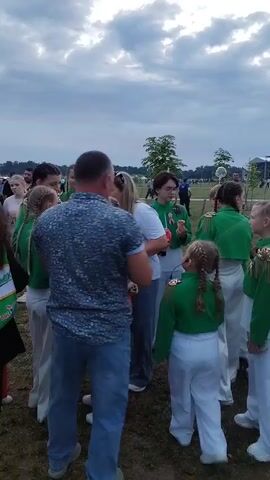 The video shows a group of people standing on a grassy area under a cloudy sky. A man in a blue patterned shirt and jeans is facing away from the camera, talking to a group of young girls in green and white uniforms. One of the girls is holding a drink. In the background, other people are walking around, some in casual clothing. There are tents and structures in the distance, suggesting an outdoor event or festival. The camera pans slightly to the left, showing more people and the tents. The focus then shifts to a man holding a smartphone and looking at it. The video is shaky and the lighting suggests it is late afternoon or early evening.