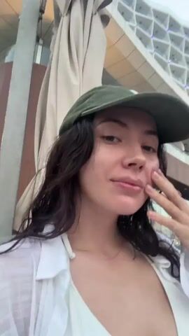 This media file features a short video clip. The first segment focuses on a young woman wearing a green baseball cap and a white bikini top with a white unbuttoned shirt over it. Her hair is dark and wavy. She is touching her face with her right hand. The background is blurred, suggesting movement, but appears to be outdoors, possibly a resort or beach setting with some structures and lighting.

The subsequent segments shift focus to the surroundings. The second segment is a very blurry shot of lights and architectural elements, likely from a building or street. The third segment presents a wider view of a clear sky with a crescent moon visible. A modern building with balconies and illuminated signage that reads "Hampton" is prominent on the right. String lights are visible in the lower portion of the frame. The overall impression is of an evening or dusk setting in a contemporary urban or resort environment. The location cues point to Ras Al-Khaimah, United Arab Emirates. There are no explicit indications of a specific event, but the imagery suggests leisure time.