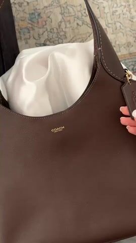 The video shows a close-up of a dark brown leather handbag. The handbag has the COACH logo in gold lettering on the front. A woman's hand with white nail polish is shown holding a small leather tag attached to the bag. The tag has gold lettering that says "NYC". The background appears to be a patterned rug. The woman's hand then moves to touch the bag.