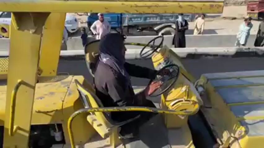 A woman in a dark garment and headscarf drives a yellow road roller on a paved road under repair. The road roller is worn, with visible signs of age and use. A group of people, mostly men in traditional clothing, stand on the side of the road, observing the operation. There are other construction vehicles and personnel visible in the background, working on the road. The environment suggests an urban or semi-urban setting in a Middle Eastern or South Asian country, with clear, bright lighting indicative of daytime.