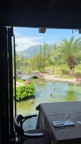 This video shows a view from a restaurant table out to a scenic outdoor area. In the foreground, a table with a dark tablecloth is set with cutlery and a glass. To the left, a dark chair is partially visible. Beyond the table, a clear, shallow stream runs through a landscaped area with lush green bushes and trees. Several ducks are swimming in the stream. A small stone bridge arches over the stream in the mid-ground. In the background, a palm tree stands to the right, and rolling green hills with mountains under a blue sky with white clouds are visible. The overall impression is a tranquil, pleasant outdoor setting.