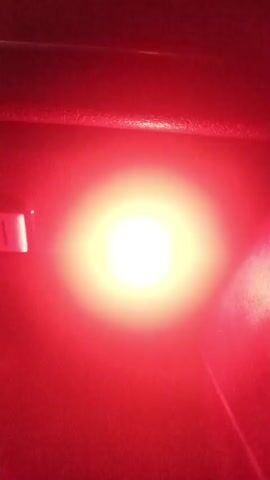 The media shows a close-up of a red flashing light. The light is round with a ribbed outer casing and a clear lens. Inside the lens, a bright red LED is visible. The light is mounted on a dark surface, possibly the interior of a vehicle or a piece of equipment. The background is predominantly dark, suggesting the scene takes place in low light conditions, perhaps at night or in an enclosed space.

The light is observed to be flashing intermittently. At certain points, the light is on, emitting a bright red glow that illuminates the surrounding area, creating a strong halo effect. At other times, the light is off, and only the red LED within the casing is visible against the dark background. The activity depicted is simply the functioning of this flashing warning light.

There are no people or animals visible in the media. The setting is dark and industrial or technical in nature. There are no clear weather or time-of-day indicators, beyond the implication of darkness. No emotions are conveyed. There is a small, rectangular white object visible on the left side of the frame in some shots, but it is indistinct. No text is legible in the media.