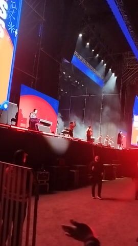 The video captures a live performance on a large stage at night, likely a concert or festival. Several performers are visible on stage, moving and dancing, enveloped in stage fog or smoke. Intense spotlights illuminate the stage, and large LED screens display vibrant colors, primarily blue and red. One screen in the bottom left corner partially shows the text "ESS". The foreground is dominated by a reddish light, where out-of-focus individuals, likely audience members, can be seen. The camera is held at an angle, looking down onto the stage and the immediate crowd area. The atmosphere appears energetic and engaging.