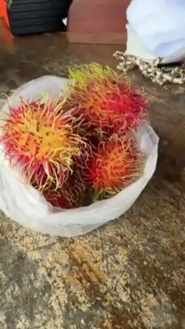 A high-angle shot depicts a plastic bag containing several rambutans on a weathered wooden table. The rambutans display a mix of red and yellow hues, with spiky, hair-like projections covering their surface. In the background, a measuring tape, a wooden cutting board, and a white towel are visible. The table's surface appears textured and mottled, hinting at exposure to the elements.