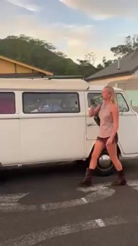 A woman with long blonde hair drinks from a green bottle while standing next to a white Volkswagen van. She is wearing a striped sleeveless top, brown shorts, and brown boots, and has a black purse on her shoulder. After drinking, she lowers the bottle and walks a few steps away from the van. The van is parked in a parking lot with trees and a house in the background, under a partially cloudy sky. The van has a white top and appears to have someone lying inside it, visible through the tinted windows.