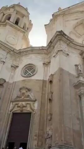 The video shows a low-angle view of the Cadiz Cathedral in Spain, panning upwards. The cathedral is a large, ornate, beige building with a bell tower, arched windows, and carved details. There are two large wooden doors, and small statues are visible on the facade. The sky transitions from overcast to sunny with light clouds. Several palm trees are visible near the cathedral, and some buildings are in the background. At the base of the building, a few people can be seen.