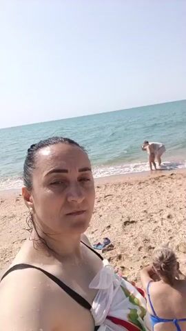 A woman in a black bikini top and a white towel wrapped around her torso stands on a sandy beach, looking at the camera. She has dark hair pulled back and is wearing earrings. Behind her, a person in white is kneeling by the water's edge, and another person in a blue bikini is sitting on the sand. The ocean is visible in the background under a clear sky. The camera pans slightly to the right, revealing more of the beach and distant beach umbrellas.