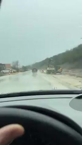 The video captures a first-person perspective from inside a moving car, looking out through the windshield on a rainy day. The road ahead is wet, and windshield wipers are actively clearing water from the glass. The steering wheel, with a Mercedes-Benz logo, is visible in the foreground, and a hand is gripping it.

On the right side of the road, there appears to be ongoing construction or roadwork. A section of the ground is unpaved and muddy, with large construction vehicles, including a yellow excavator and a dump truck, present. Orange and white traffic barriers or cones mark the edge of this construction zone.

Further down the road, other cars are driving, their headlights and taillights creating reflections on the wet asphalt. The surrounding environment consists of a hilly or mountainous terrain in the distance, partially covered with sparse vegetation. The sky is overcast and grey, indicating the rainy weather. The sound of the car's engine, road noise, and the distinct sweeping sound of the windshield wipers are audible throughout the clip.