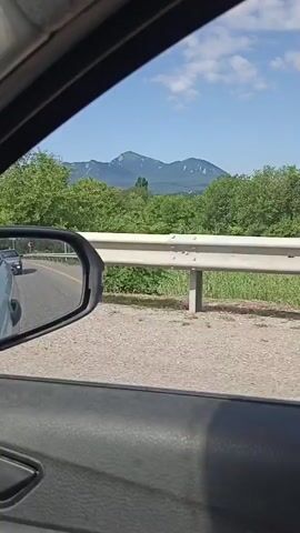 The video is shot from inside a moving vehicle, likely a car, looking out the passenger side window. The perspective shows the road, a guardrail, and lush green trees and vegetation. In the distance, a mountain range is visible under a partly cloudy blue sky. A road sign with red and white markings is also present. The driver's side mirror reflects the road and passing vehicles. The interior of the car, including a portion of the door panel, is also visible in the foreground. The overall impression is a scenic drive through a rural or mountainous area.