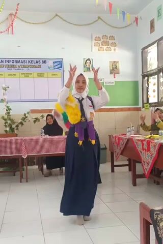 A young female student, wearing a white hijab, a white shirt with a school uniform tie, and a dark blue skirt, is performing on a stage decorated with colorful triangle flags. She is holding yellow pom-poms and appears to be dancing. In the background, other students and adults are visible, some seated at tables covered with red and white checkered tablecloths. The room looks like a classroom or an event space, with a whiteboard or bulletin board on the wall displaying information. The lighting is bright, suggesting an indoor setting with artificial lights. The video is recorded vertically.