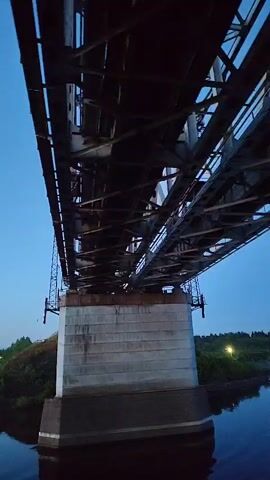 The video shows a low-angle view of a steel truss railroad bridge spanning a body of water. The bridge is supported by concrete piers. Vegetation lines the shore on both sides of the river.  The sky is a gradient of dark blue to light blue, indicating either early morning or late evening. A single light is visible on the bridge structure. The water is calm, reflecting the bridge and sky.  No people or other specific actions are visible in the frame.