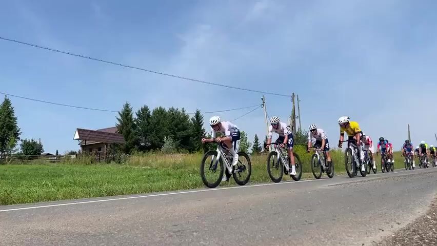 A group of cyclists are racing on a paved road on a sunny day. The cyclists are wearing brightly colored jerseys and helmets, and they are riding racing bicycles. The road is lined with green grass and trees, and there are power lines running overhead. The cyclists are moving from left to right in the frame. The camera is positioned low to the ground, giving the impression of being close to the action. The lead cyclist is wearing a yellow and green jersey and is slightly ahead of the rest of the group. Another cyclist in a blue jersey is slightly behind the lead cyclist. The remaining cyclists are in a tight pack behind them. The sky is blue with a few white clouds.