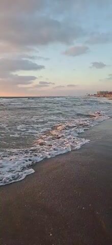 A slow-motion video shows waves breaking on a sandy beach. The sky is overcast with soft pink and blue clouds, suggesting sunrise or sunset. The water is choppy and appears somewhat murky. There are no people or distinct objects visible in the foreground or background, only the natural environment of the ocean and the beach.