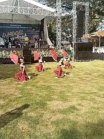 The video shows five young girls performing a traditional dance on a grassy area. They are wearing matching red and gold costumes and each holds a red fan. In the background, there is a stage with a band performing and a banner that reads "Pentas Seni Dalam Rangka HUT Kab. Sragen Ke-280". There are also large speakers and lighting trusses around the stage. The sky is partly cloudy.