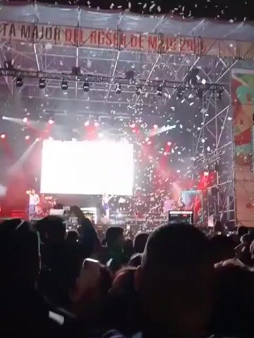 The video shows a live concert at night. Onstage are several performers wearing colorful outfits. Above them is a large screen projecting bright light. The stage is decorated with a metal truss structure. Confetti is falling from above. A crowd of people is visible in front of the stage, many holding up their phones to record the performance. The stage has lighting fixtures, including red spotlights, directed toward the audience. A banner at the top of the stage is visible. The overall atmosphere is lively and celebratory.