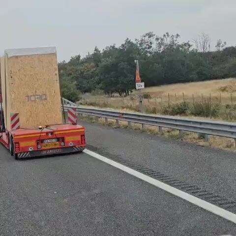 A red truck is transporting a large, light-colored rectangular object on a flatbed trailer. The object appears to be made of plywood and has the word "TORO" printed on its side. The truck is on a highway, indicated by the road markings and a guardrail. In the background, there are trees and a cloudy sky. There is also a sign overhead with Italian text and numbers. The truck and its cargo are moving forward.