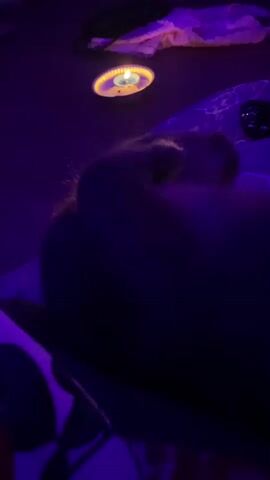 The video shows a dark room lit by a candle. A hand is caressing a dark-haired person lying down. There is a cloth visible on the right and a patterned surface behind the person’s head. The video is in low resolution and very dark.