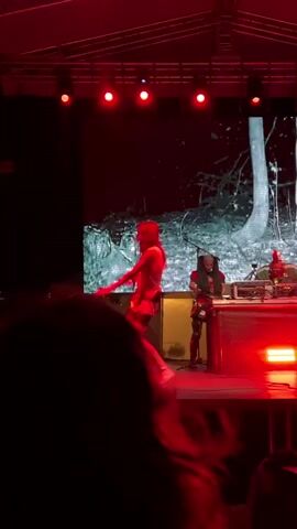 A person dances on a stage under red lights. The person is wearing a dress and boots. Behind them is a large screen showing an image of trees. There are speakers and other equipment on the stage. In the background, people are watching and taking photos with their phones.
