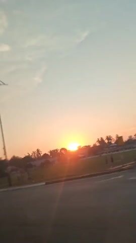 The video shows a sunset over a road. A street light and a bus stop shelter are visible on the left side of the road. The sun is setting behind trees in the distance, casting an orange glow across the sky. The sky is partly cloudy. The road is paved and has white lines marking lanes. The camera is moving, likely from inside a vehicle, giving a sense of travel. There are no people clearly visible in the video. The environment appears to be a semi-urban or suburban area with some greenery.