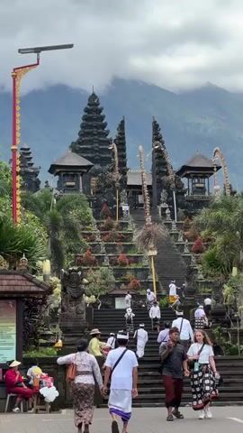 The video shows a large, tiered Balinese temple complex with a mountain in the background. The temple is made of dark stone and features multiple levels with stairs leading up to the top. The steps and platforms are adorned with plants and decorations. People are walking up and down the stairs, some wearing traditional Balinese clothing. The sky is overcast, and clouds partially obscure the mountain in the background. A tall red and gold lamppost stands to the side of the temple. Signage is visible at the bottom of the steps.