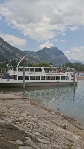 The video opens with a view of a white boat named "Quintin" docked at a pier on a lake. The boat has passengers on its upper deck. Mountains rise in the background under a cloudy sky. The camera then pans across the lake, showing calm water and distant trees. The final shots focus on a fountain in the middle of the lake, spraying water upwards, with the mountains and trees visible behind it. The shoreline in the foreground consists of rocks and sand.