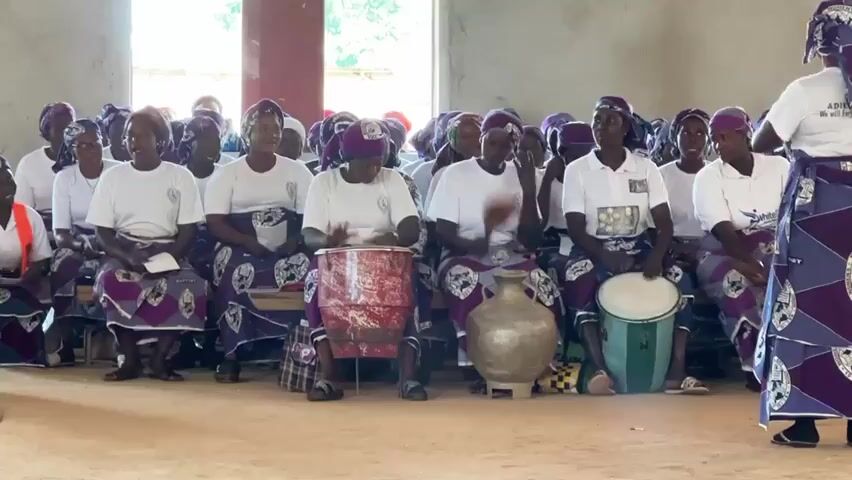 The image depicts a group of women, likely participating in a cultural or religious gathering. They are seated in rows, many of them wearing matching white t-shirts and vibrant purple and white patterned skirts and headscarves. Some women are playing drums, with one woman in the foreground actively drumming a red and white cylindrical drum, while another plays a white drum to her right. A large, metallic pot or vase sits between two drummers.

In the background, more women are seated, all similarly dressed, suggesting a unified group. To the far right, a woman stands with her arms raised, seemingly leading or directing the group, also dressed in a similar outfit. The overall mood appears energetic and celebratory.

The setting seems to be an indoor space with plain, light-colored walls and natural light coming from what appears to be an open window or doorway on the left, casting shadows on the floor. The floor is a reddish-brown dirt or sand. The time of day appears to be daytime due to the natural light. The presence of musical instruments and the spirited posture of the woman leading suggest a performance, rehearsal, or ritual.

Visible text on the t-shirt of the woman leading reads "ADIDA DA" and below it "WE WILL ADVISE OUR DAUGHTERS." This text hints at the group's identity and possibly the message they are conveying. The clothing and the drumming suggest a West African cultural context, consistent with the provided location of Kufana, Nigeria. The emotions conveyed are positive, with a sense of community and shared activity.