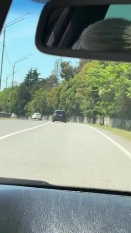 The video provides a first-person perspective from inside a moving car, looking forward through the windshield on a sunny day. The lower part of the car's dashboard is visible at the bottom of the frame. The rearview mirror reflects the top of a person's head, showing light-colored hair, likely blonde or grey.

The scene depicts a clear, multi-lane road, possibly a highway, with white solid and dashed lines marking the lanes and edges. Several other vehicles are visible ahead, including a dark-colored SUV or minivan directly in front, followed by two white cars and a red truck further down the road. The car appears to be following the flow of traffic.

Both sides of the road are lined with dense, lush green trees and foliage, suggesting a verdant, possibly hilly or mountainous landscape. On the right side of the road, a low stone or concrete wall separates the roadway from the natural vegetation. Utility poles are visible on the left side of the road, paralleling the asphalt.

The sky is bright and clear blue, indicating fair weather and bright sunshine. The time of day appears to be midday or early afternoon. The overall environment suggests driving through a scenic, natural area. The location context indicates this is Primorskoe, Abkhazia.

Background music, sounding like pop or electronic with a noticeable beat, is playing throughout the video. No visible text can be discerned in the recording.