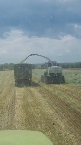 The video shows a Claas Jaguar forage harvester cutting and chopping crop in a field and ejecting it into a trailer pulled by another vehicle driving alongside it. The sky is partially cloudy. The field has already been partially harvested.